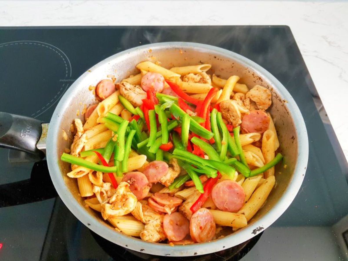 bell peppers, pasta, smoked sausage, chicken pieces and onions cooking in a skillet.