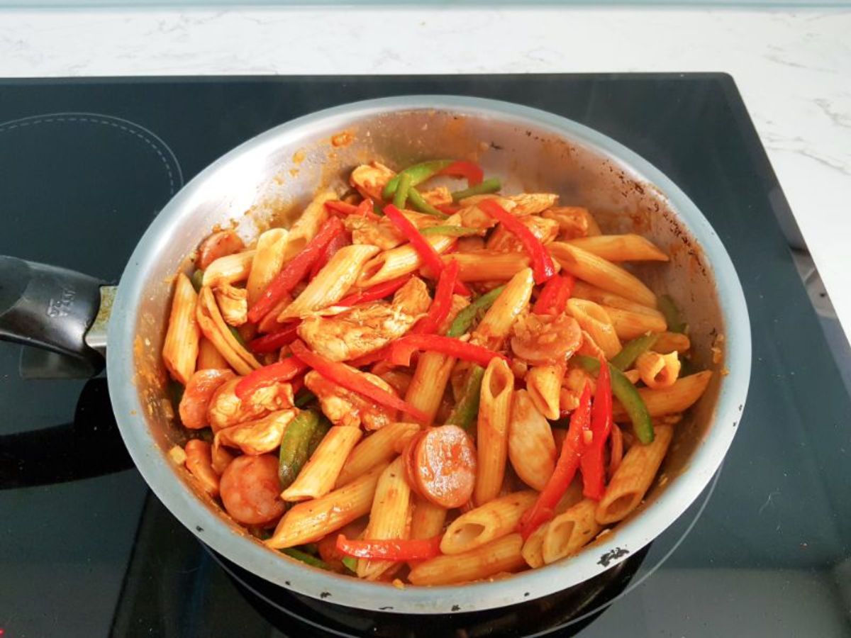 bell peppers, pasta, smoked sausage, chicken pieces and onions cooking in a skillet.