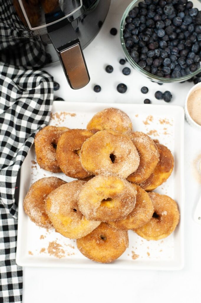 Overhead view of a plate of cinnamon sugar donuts next to black and white checked dish cloth and below an air fryer and bowl of blueberries