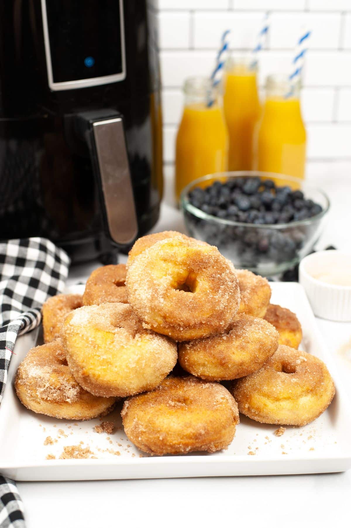 Plate of cinnamon sugar air fryer biscuit donuts in front of bowl of blueberries, glasses of orange juice and an air fryer
