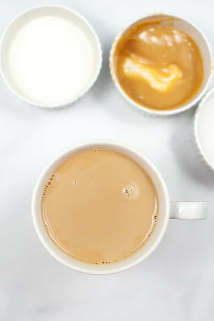 coffee with creamer stirred in, in a coffee cup, next to sugar and caramel sauce in white bowls