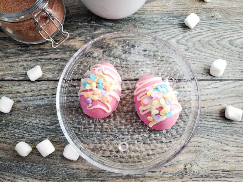 two Easter egg hot cocoa bombs on a glass plate surrounded by marshmallows on a wood table next to a white cup of hot chocolate and cocoa in an open glass jar