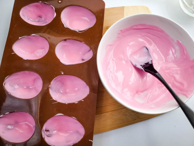 pink melted candy in a white bowl with a black spoon next to an egg mold coated with melted pink candy