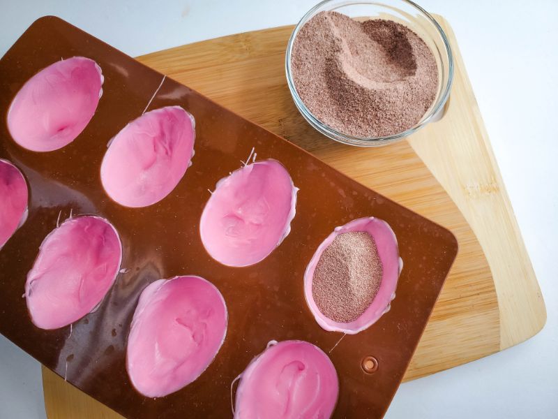 an egg mold coated with melted pink candy being filled with cocoa powder