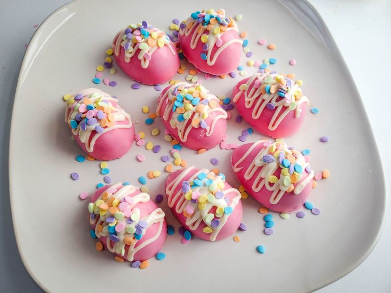 pink hot cocoa bombs with drizzled white frosting and sprinkles on a white plate