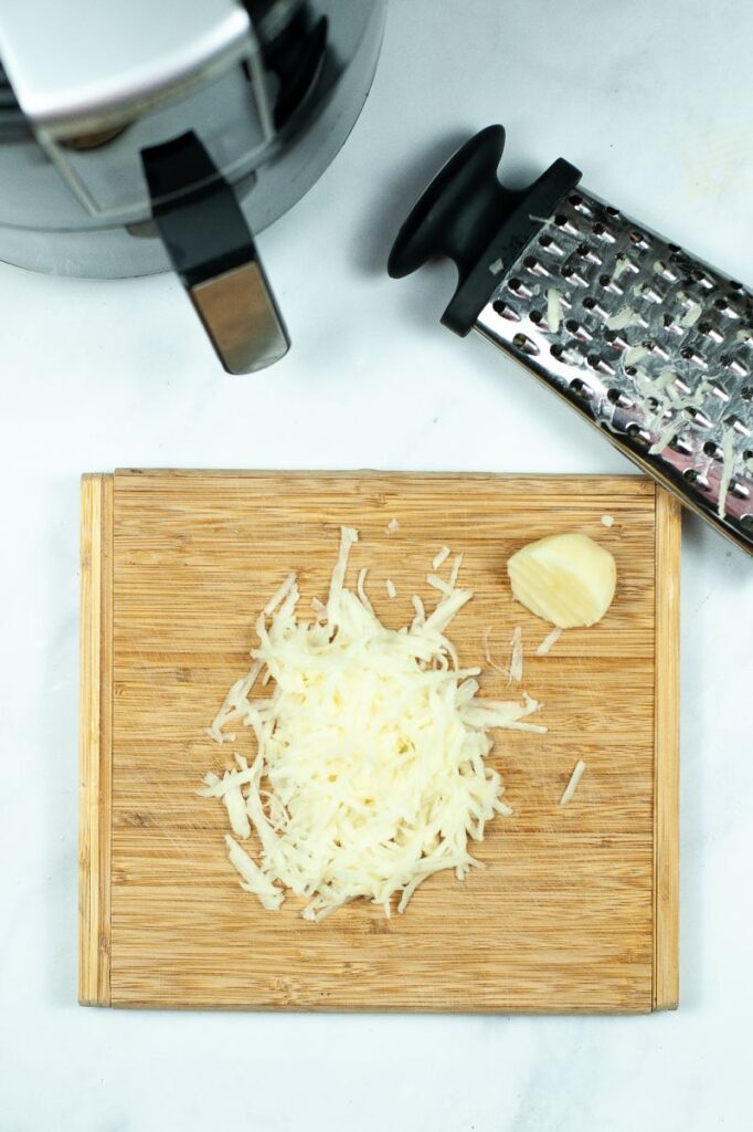 shredded potatoes and half a potato on a wooden cutting board next to a cheese grater and an air fryer on a white table