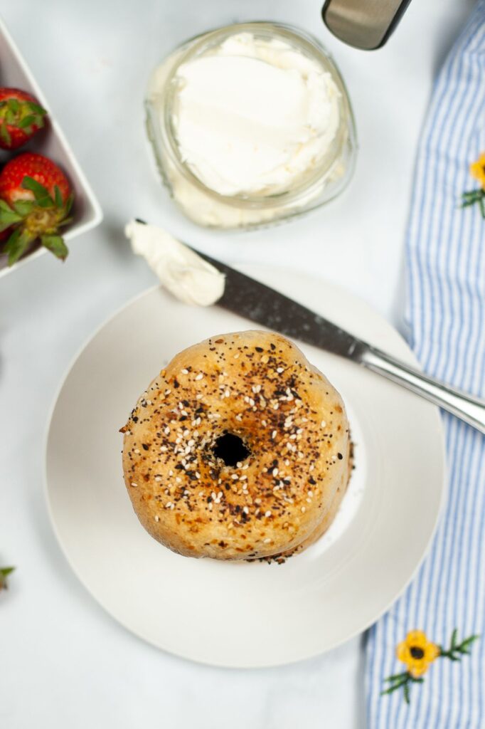 overhead view of a bagel and a knife with cream cheese on it, on a white plate, next to a jar of cream cheese and some strawberries
