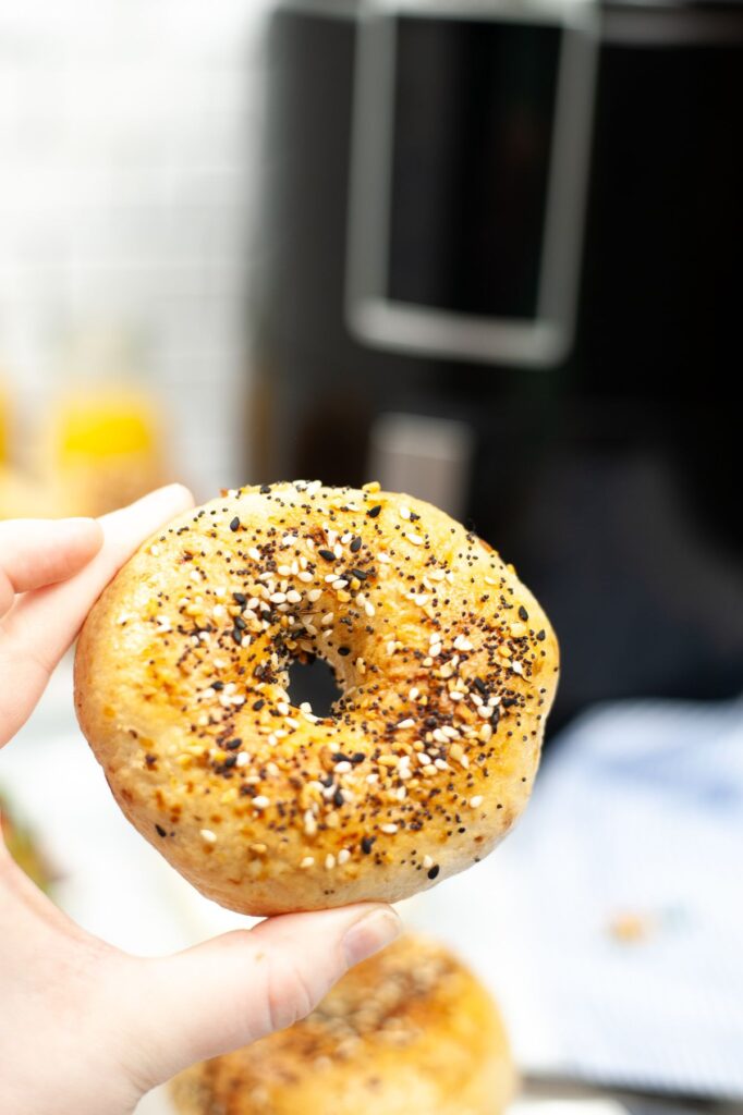 a hand holding a seasoned bagel with an air fryer in the background