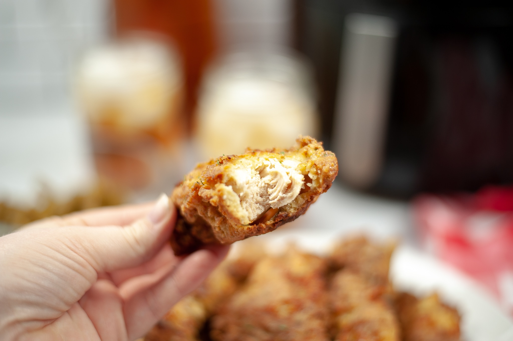 close up of a hand holding an Air Fryer Chicken Tender with a bite taken out of it, with a plate of Air Fryer Chicken Tenders blurred in the background