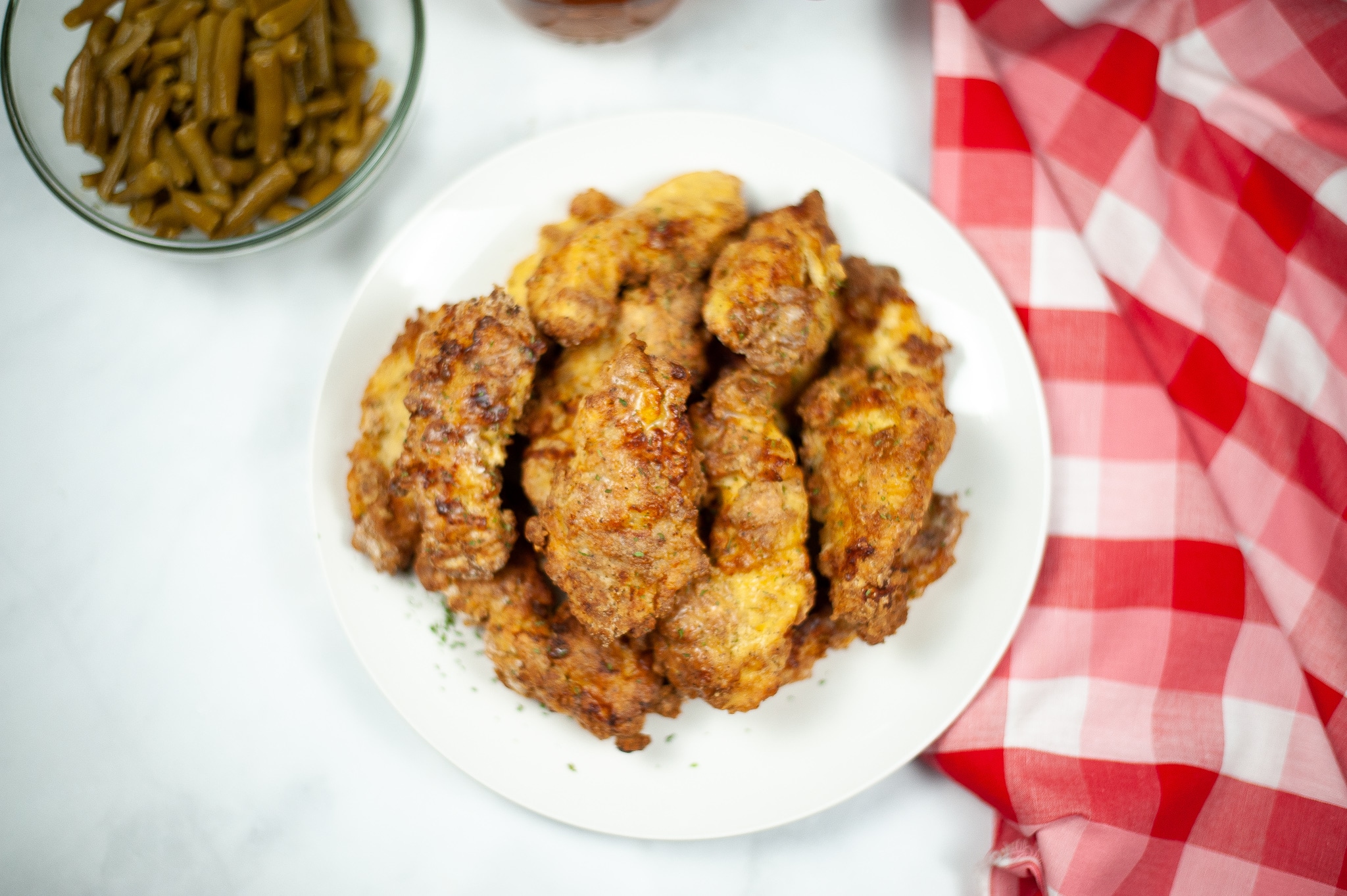 closeup overhead view of Air Fryer Chicken Tenders on a white plate next to a red and white checkered cloth and green beans in a glass bowl