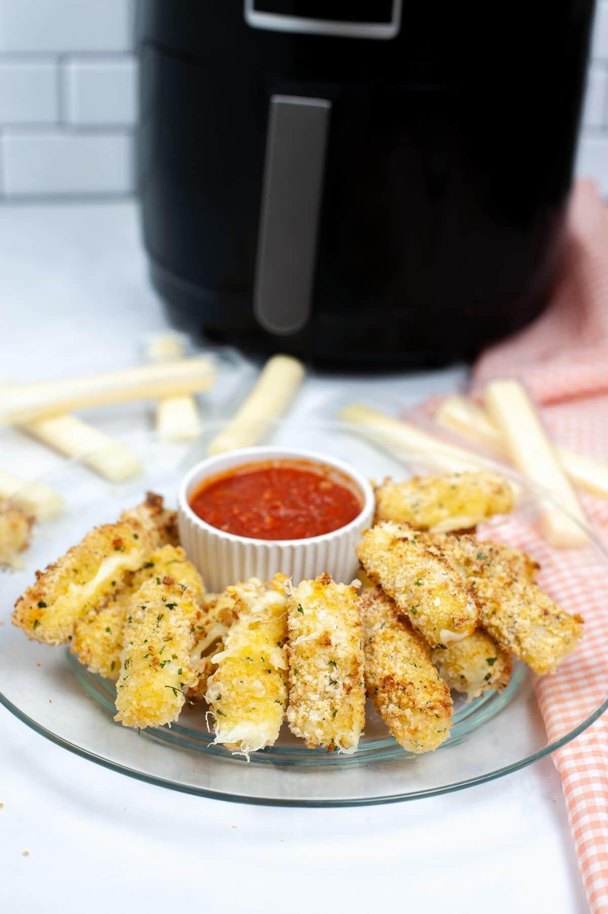 Air Fryer Mozzarella Sticks on a glass plate with a sauce in the middle and air fryer in the background.