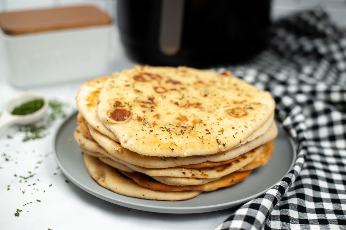 A horizontal image of stacked Air Fryer Naan on a gray plate with blurred tablespoon of parsley garnish and air fryer on the background.