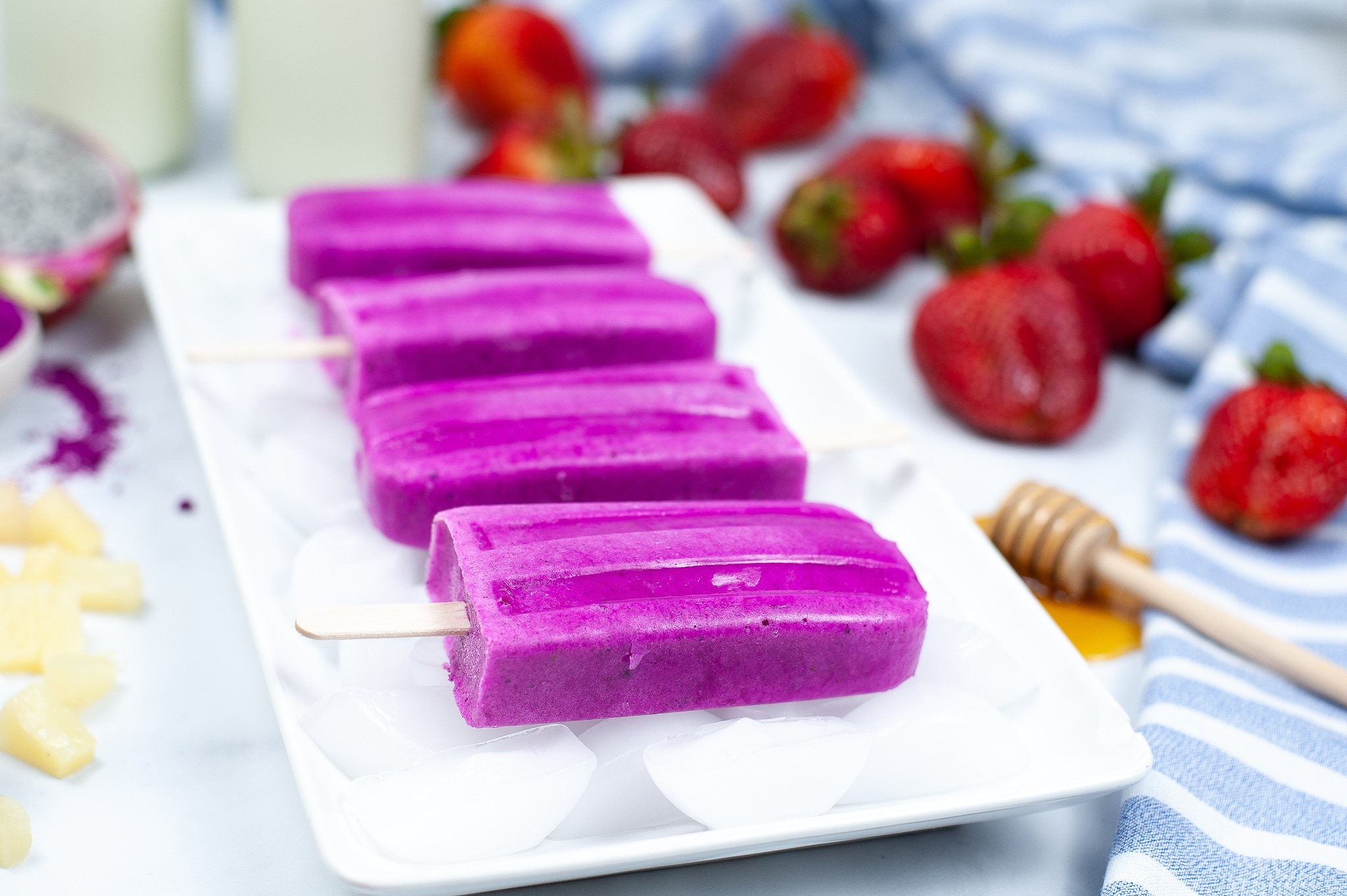 dragon fruit popsicles on top of ice cubes on a white plate with strawberries in the background