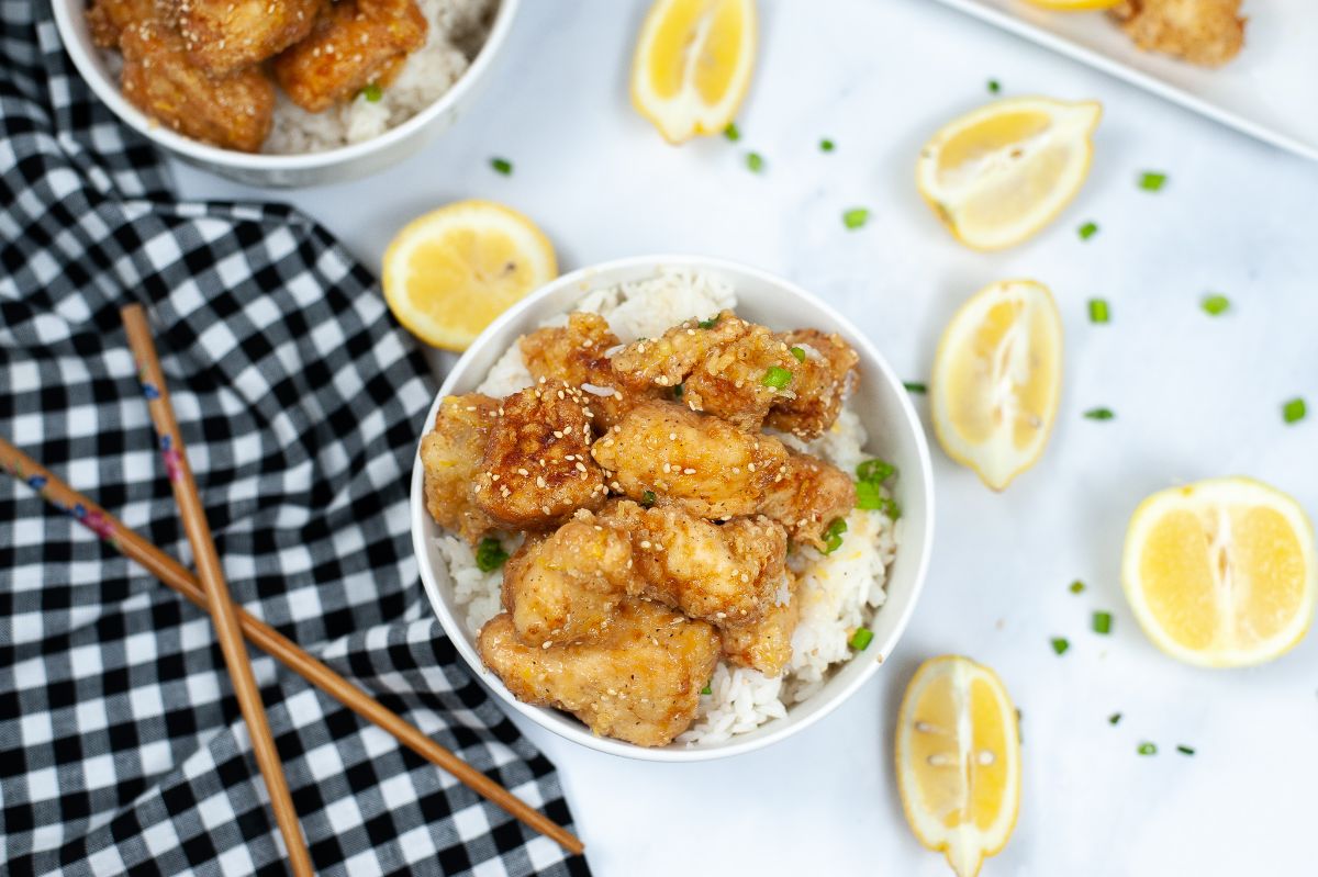 overhead view of lemon chicken and white rice in a white bowl next to sliced lemons, chopsticks and a black and white checkered cloth