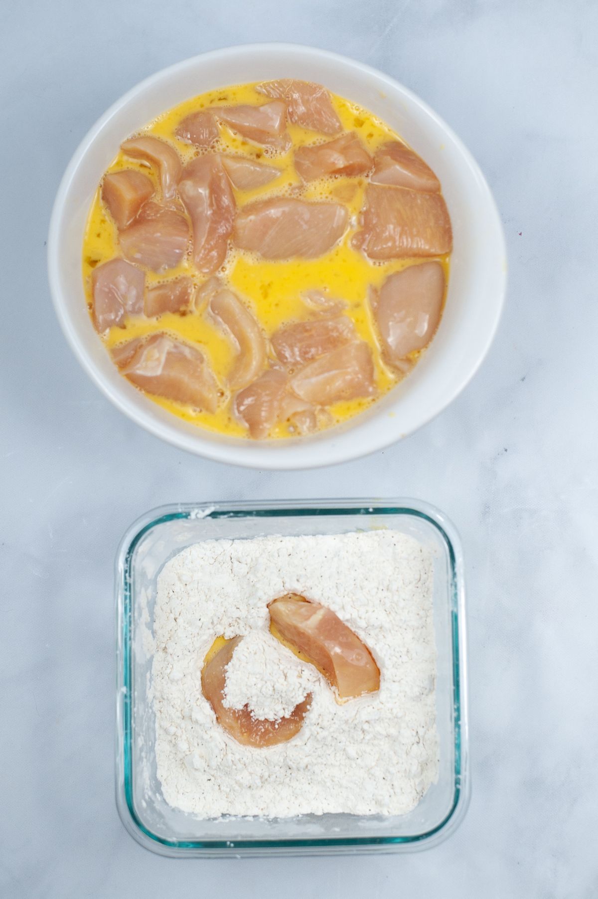 raw chicken in a bowl of eggs next to a glass dish with raw chicken being coated in flour