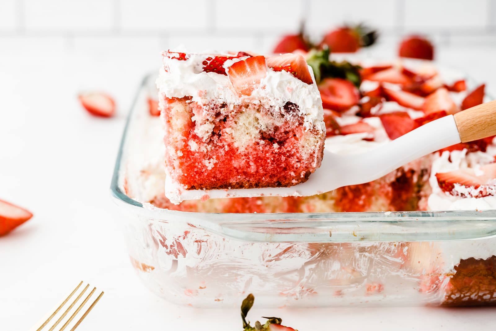 slice of strawberry poke cake on a spatula above the rest of the cake in a glass dish