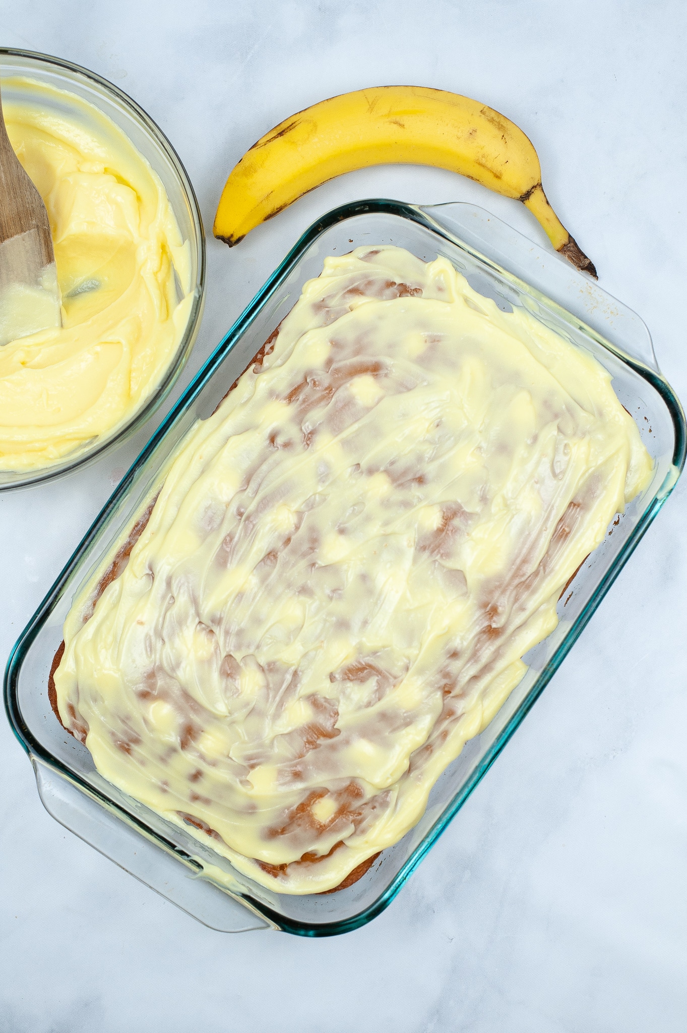 banana poke cake with pudding on top next to a banana and more pudding mixture in a glass bowl