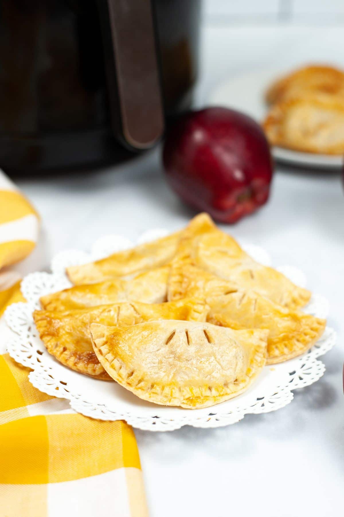 A vertical image of Air Fryer Caramel Apple Empanadas on a white serving plate with a blurred air fryer and apple in the background