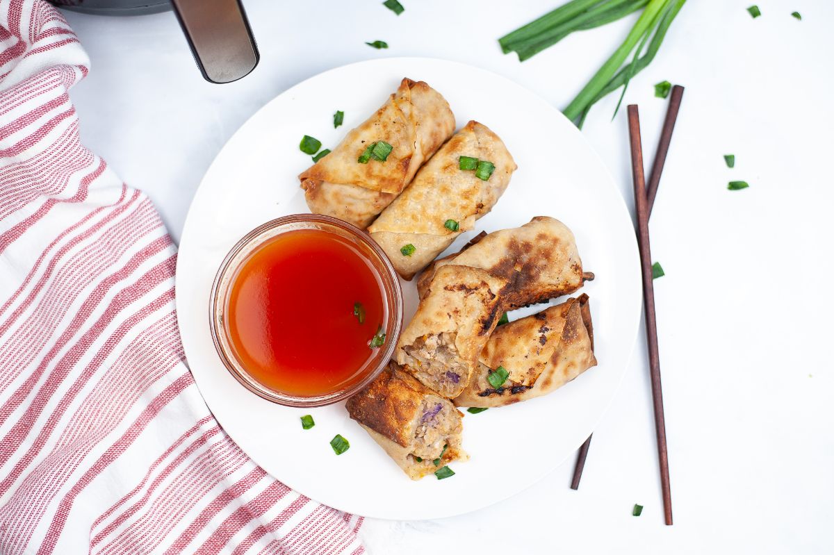 A horizontal overhead image of Air Fryer Chicken Egg Rolls on a white serving plate with egg roll sauce and a a pair of chopsticks beside the white plate.
