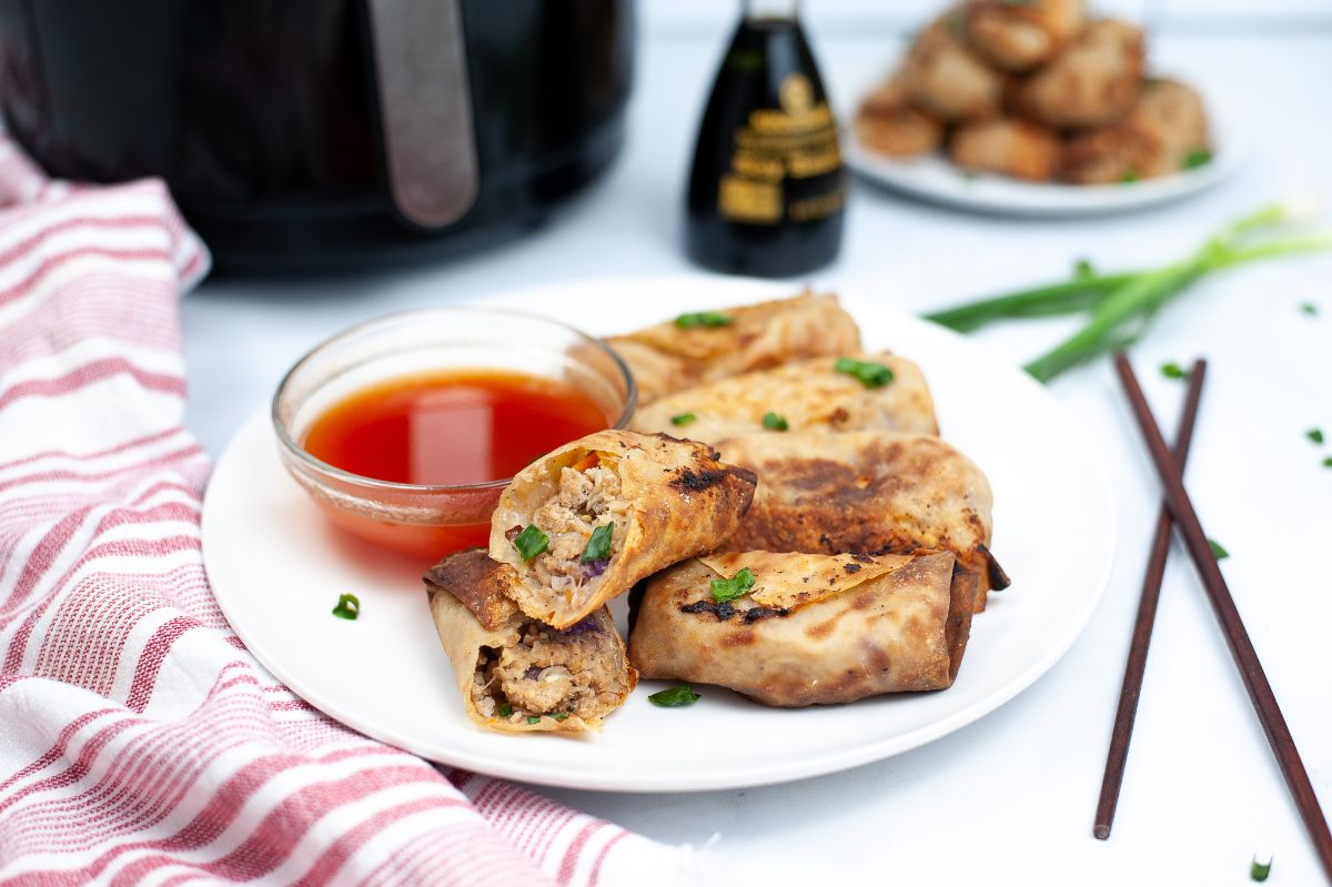 A horizontal image of Air Fryer Chicken Egg Rolls on a white serving plate, with egg roll sauce, and with one egg roll halved to show the meat inside.