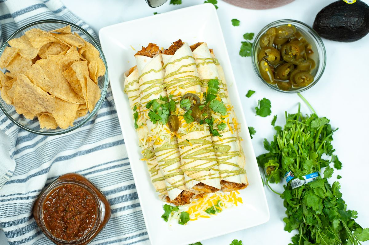 Air Fryer Chicken Taquitos stacked on a white rectangular serving plate with nachos, jalapenos, salsa around the plate.