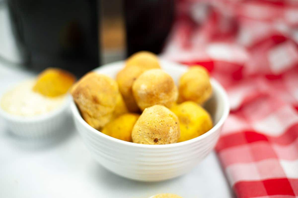 Air Fryer Hush Puppies in a white serving bowl with an air fryer, dipping sauce and a red and white checkered cloth blurred in the background.