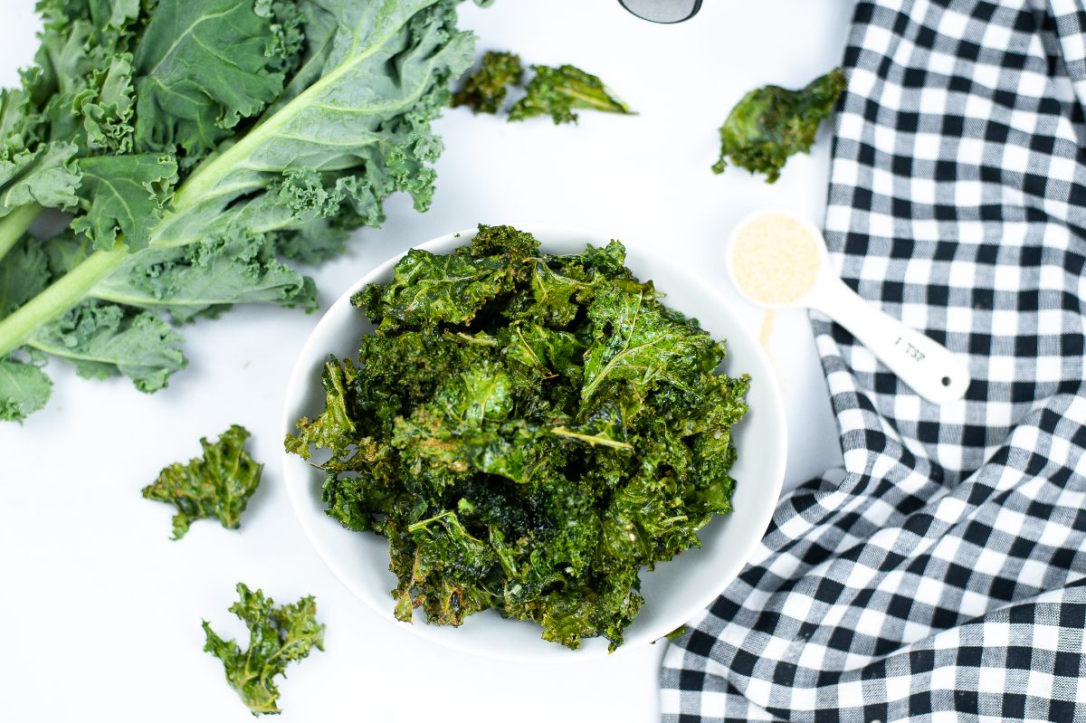 A horizontal overhead shot of Air Fryer Kale Chips in a bowl with measuring spoon on the right side and fresh Kale on the left.