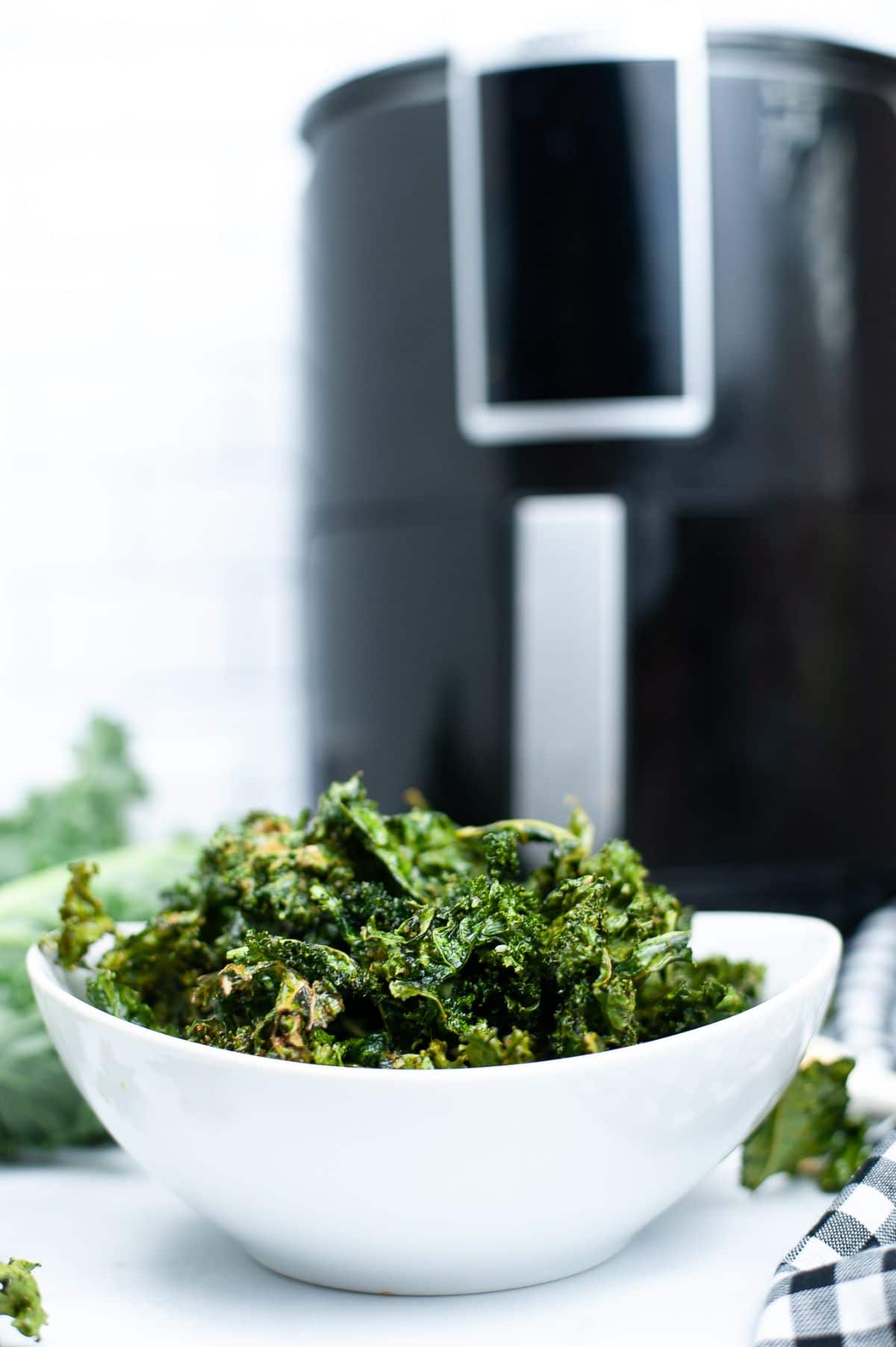 A vertical shot of the Air Fryer Kale chips in a white bowl with a blurred air fryer in the background.