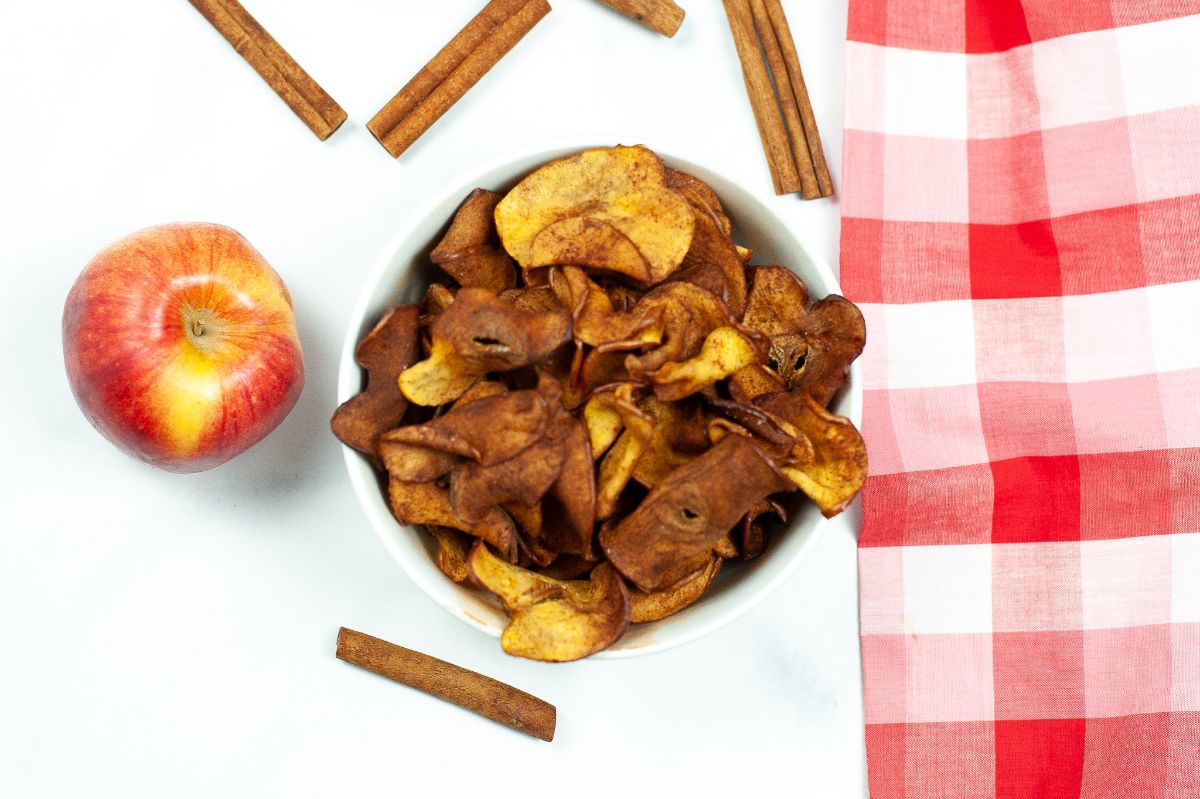 A horizontal overhead shot of Air Fryer Cinnamon Apple Chips in a white bowl with an apple and some cinnamon sticks surrounding the bowl.