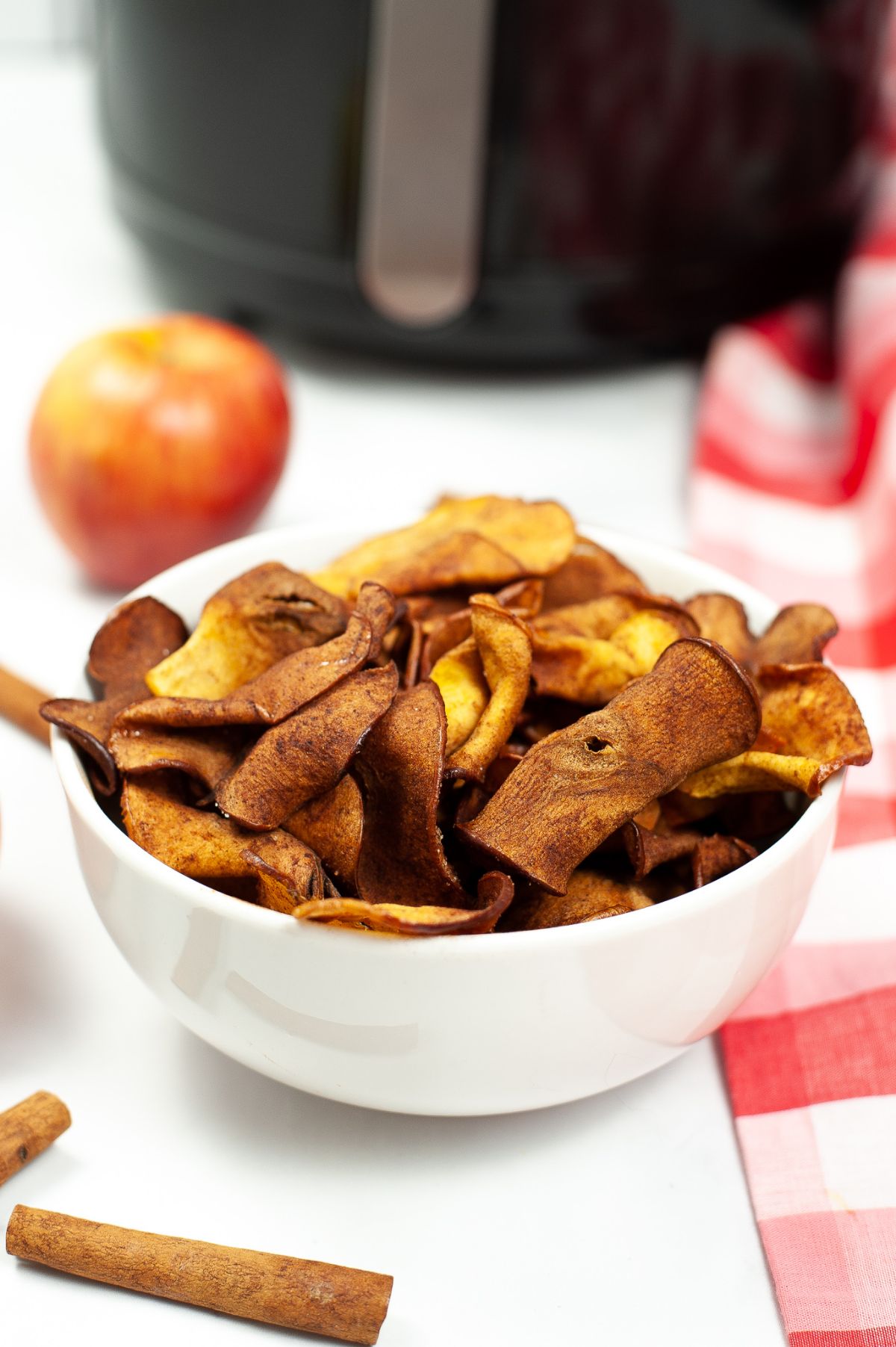 A vertical image of Air Fryer Cinnamon Apple Chips in a white bowl with an apple and an air fryer in the background