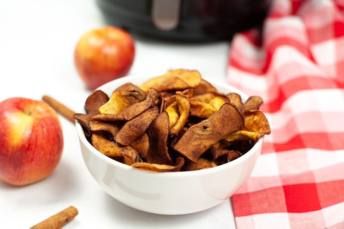 A horizontal image of Air Fryer Cinnamon Apple Chips in a white bowl next to apples and a red and white checkered cloth