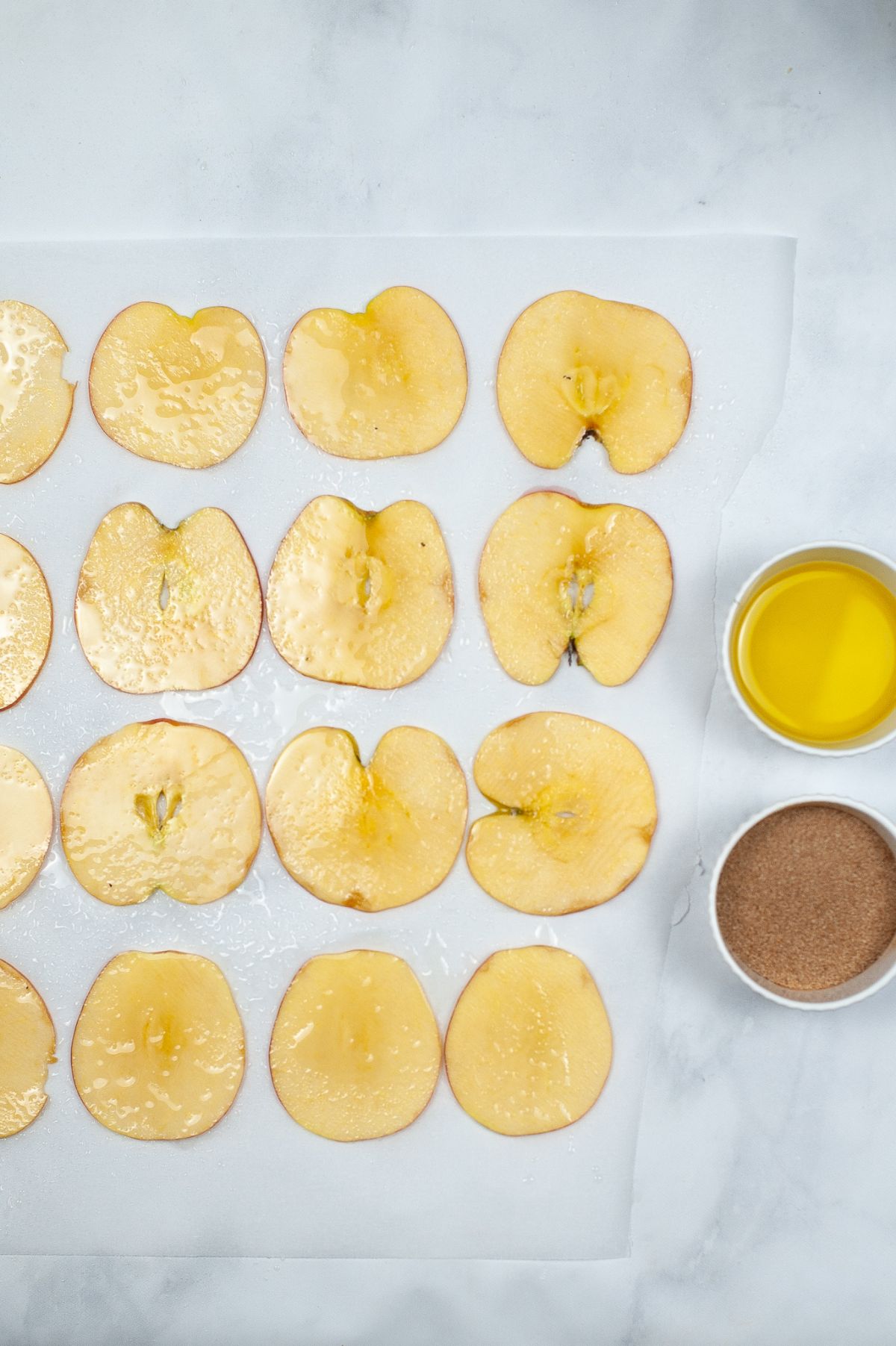 Thin slices of apple on a parchment paper next to bowls of oil and cinnamon