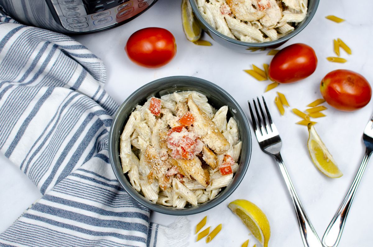 An overhead shot of Instant Pot Cajun Chicken Pasta in a bowl with a fork and sliced lemon on the right side; another bowl, Instant Pot, and raw tomatoes above it.