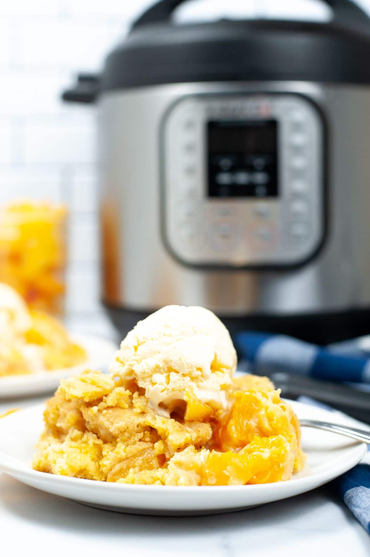 A vertical image of Instant Peach Cobbler topped with a scoop of ice cream on a white serving plate, shot from the side to emphasize the layers of the peach cobbler.