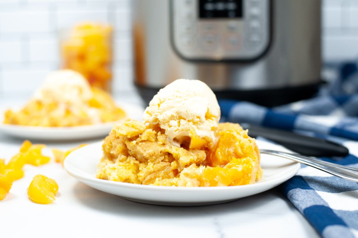 A horizontal image of Instant Peach Cobbler topped with a scoop of ice cream on a white serving plate, shot from the side to emphasize the layers of the peach cobbler.