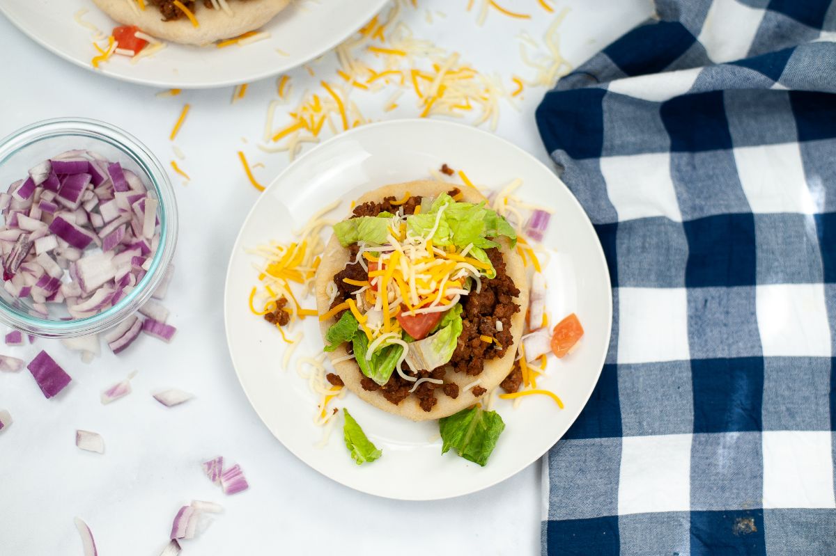 A horizontal overhead shot of Air Fryer Indian Bread Tacos on a white plate with a generous amount of taco meat and other taco ingredients on top of it with onions and cheese on the table beside the plate.