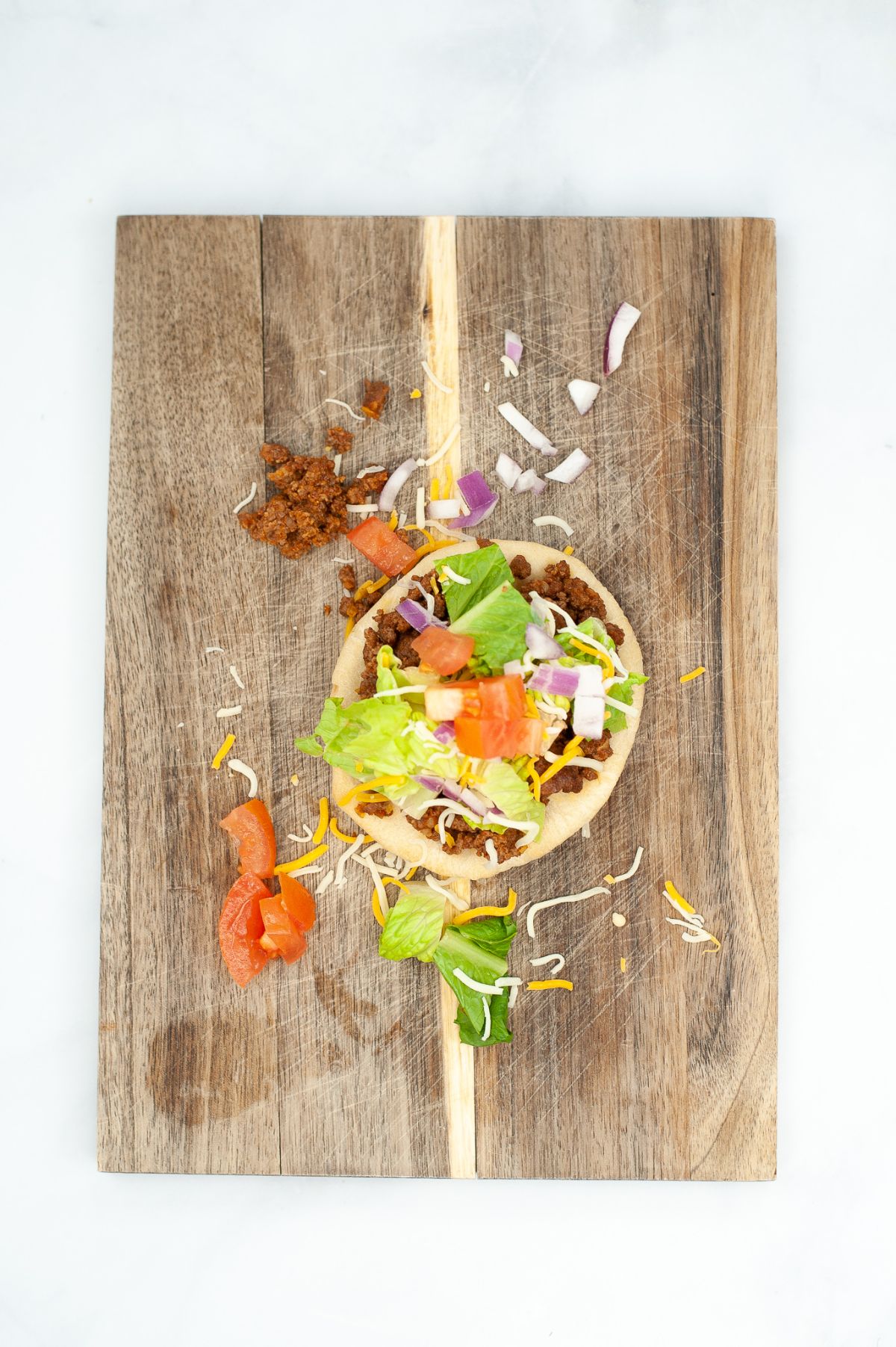 Taco fry bread on a wooden chopping board being topped with taco meat, lettuce, cheese, onion, and tomatoes.