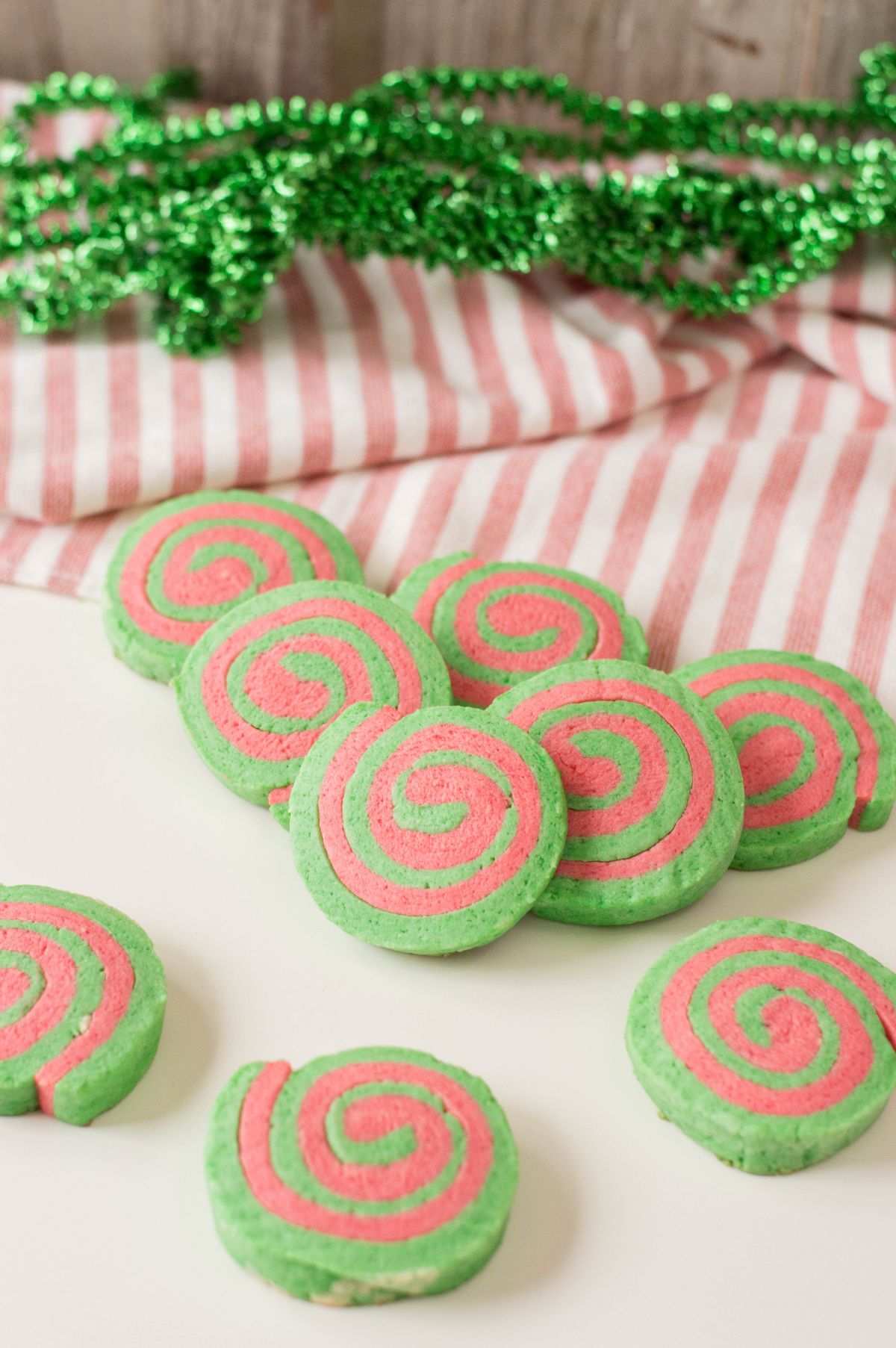 Vertical image of Christmas Pinwheel Cookies scattered on the table with green garlands above it and a table cloth with white and red stripes.
