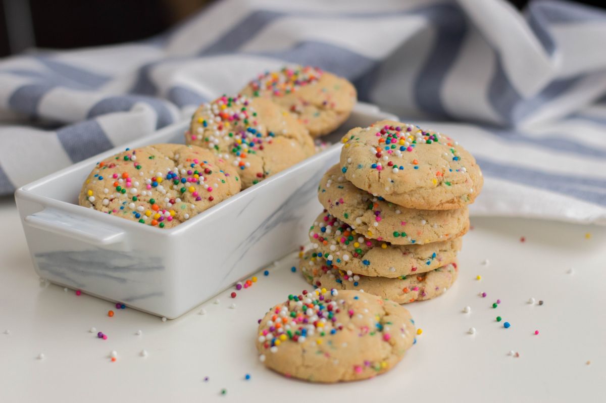 A vertical image of Funfetti cookies, a stack of 4 cookies, a single cookie in front of the stack, and other cookies in  rectangular ceramic serving dish behind the stack.