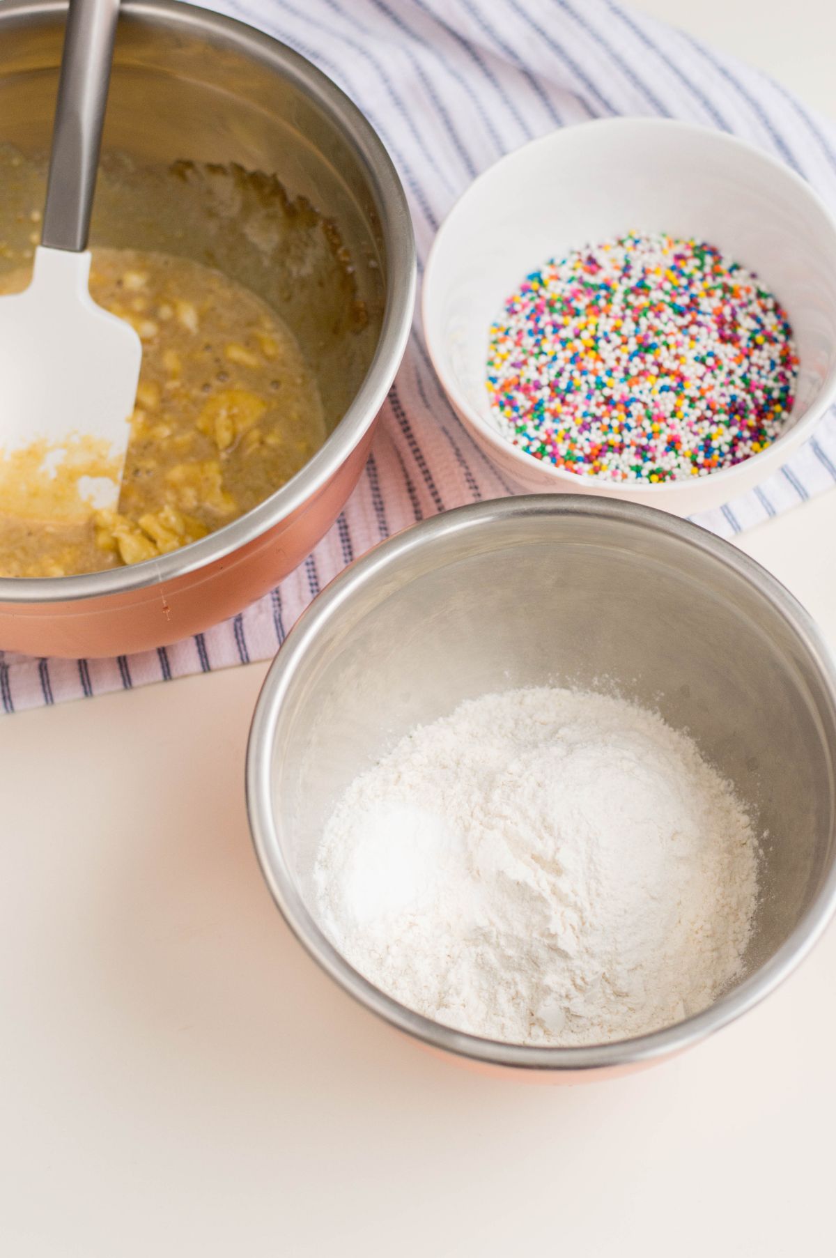 Ingredients used to make Funfetti cookies, the wet ingredients combined in a large bowl, dry ingredients in a smaller bowl, and the sprinkles in another smaller bowl.
