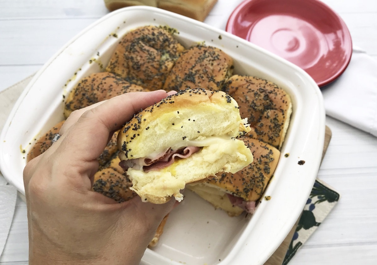 a hand holding a ham and cheese slider above more sliders in a white dish.