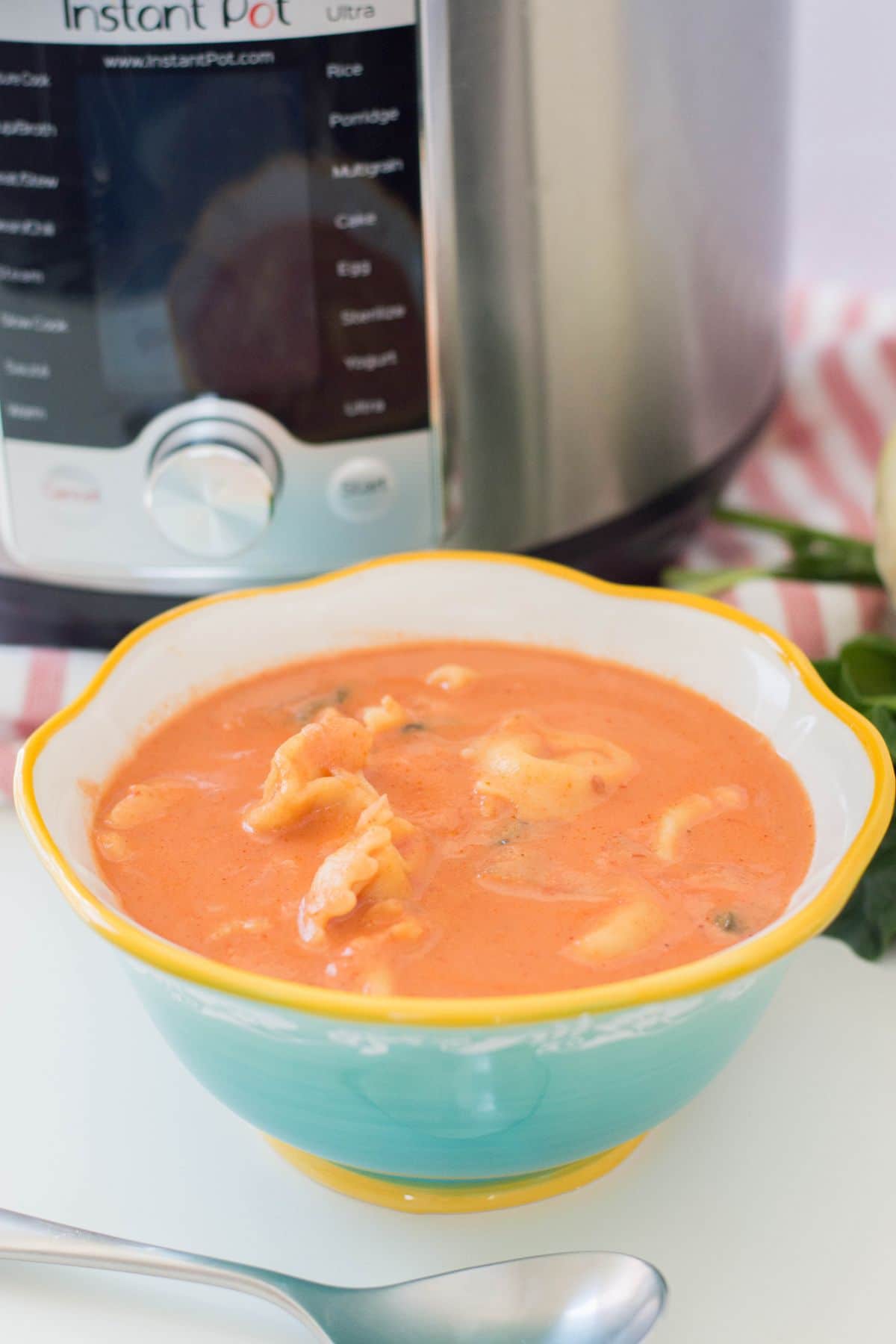 A vertical image of Instant Pot Tortellini Soup in a bowl with a spoon below it and instant pot in the background.