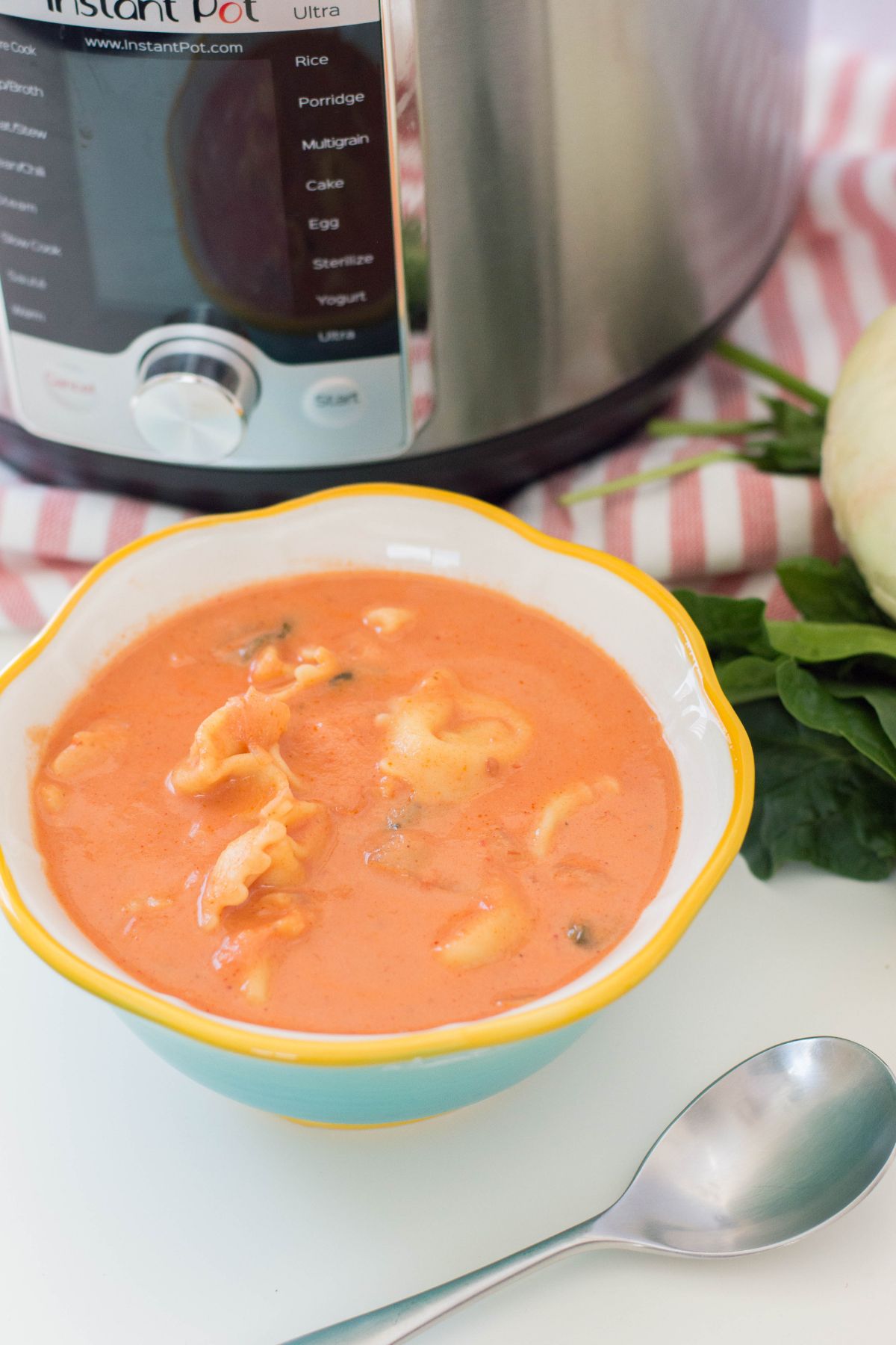 A vertical image of Instant Pot Tortellini Soup in a bowl with a spoon beside it and instant pot in the background.