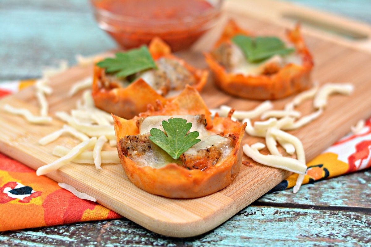 mini meatball appetizer cups next to shredded cheese on a wooden cutting board.