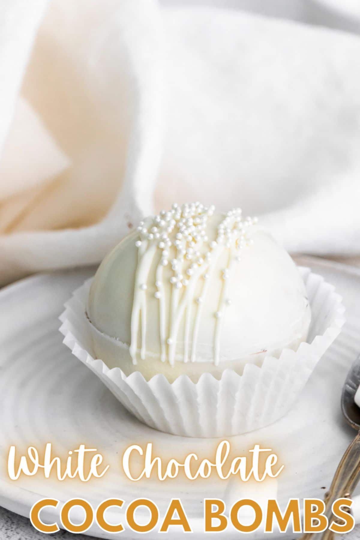 A vertical image of a White Chocolate Hot Cocoa Bomb on a white serving plate with a spoon beside it and cursive text at the bottom saying "White Chocolate Cocoa Bombs"