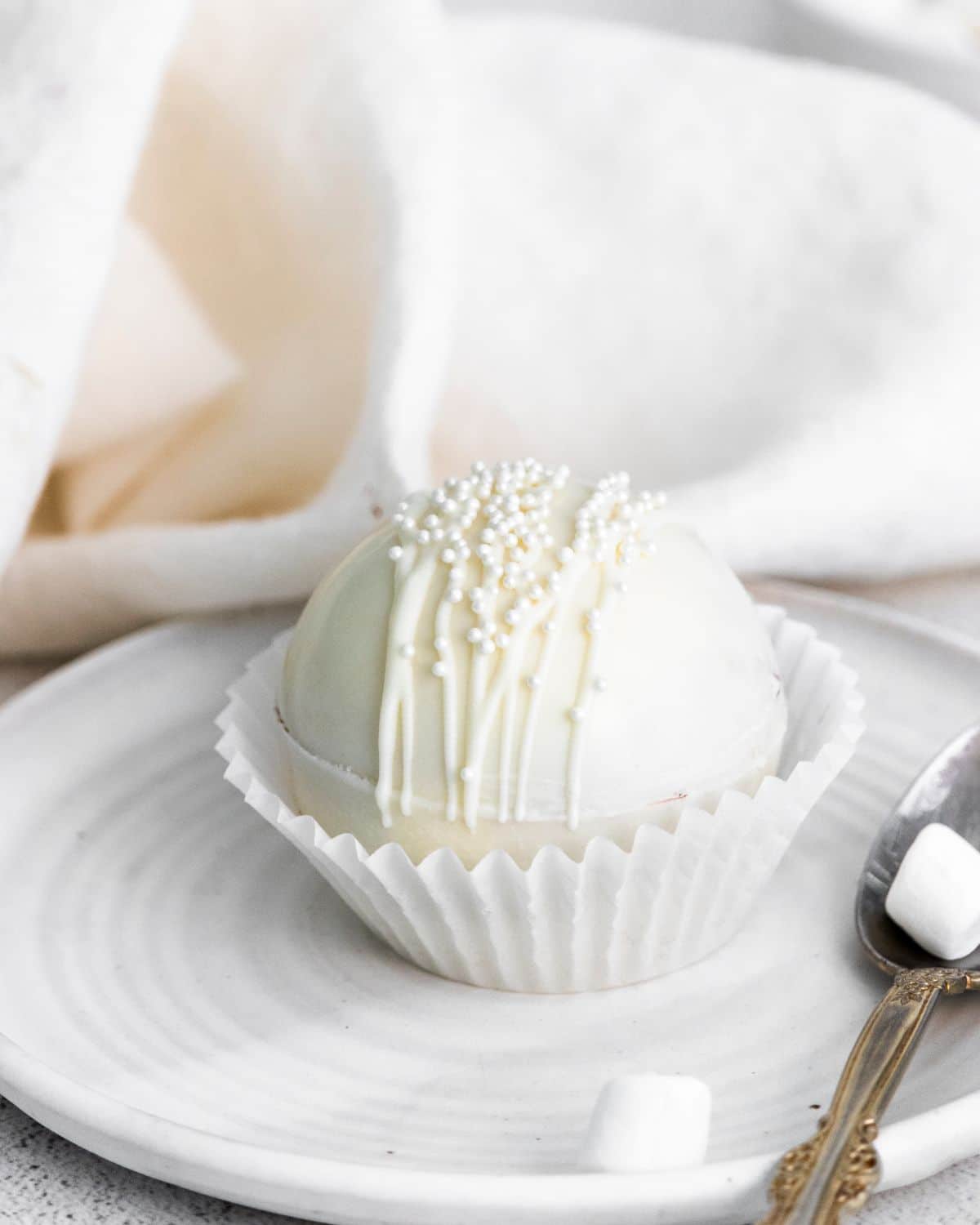 A vertical image of a White Chocolate Hot Cocoa Bomb on a white serving plate with a spoon beside it.