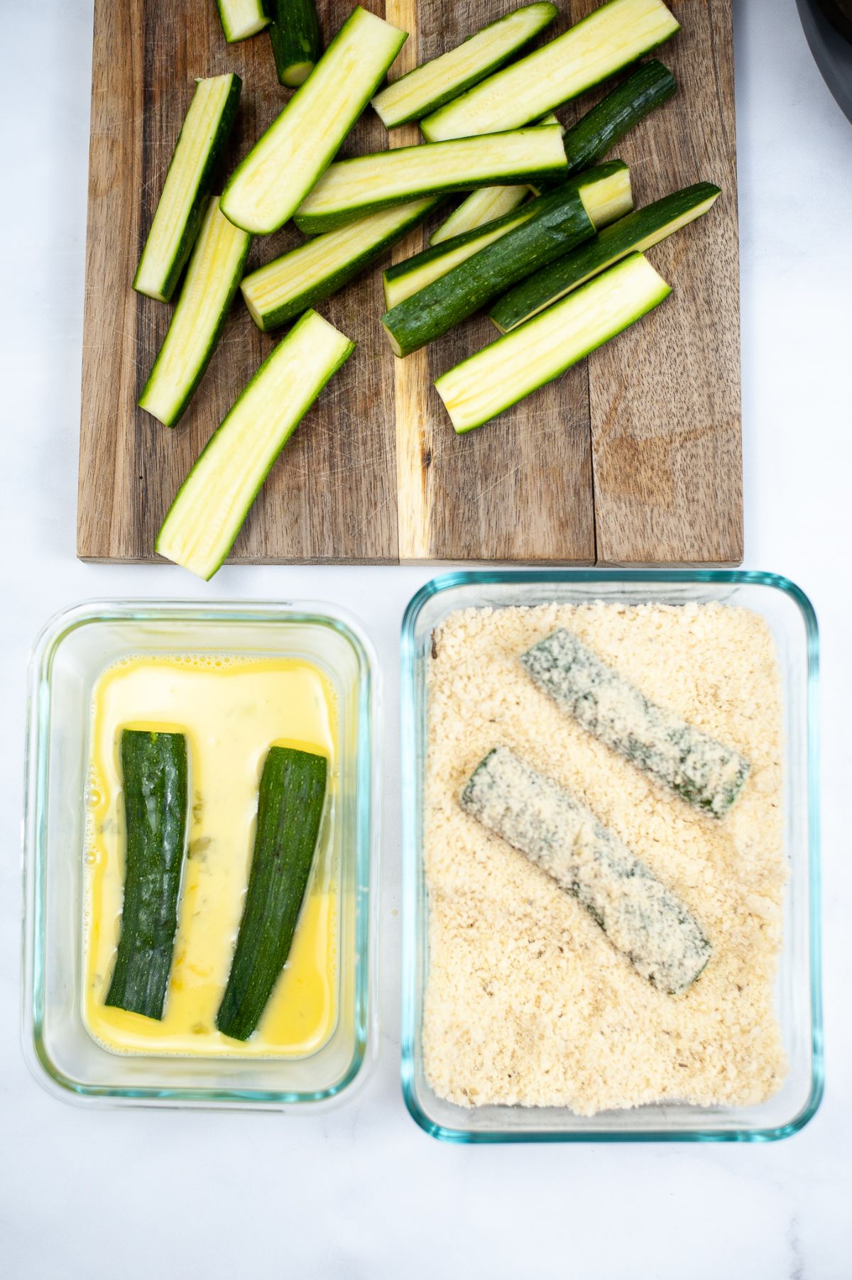 An image showing the first 3 steps in making Air Fryer Zucchini Fries. Sliced Zucchinis on a wooden chopping board, two Zucchinis dredged in eggs, and another two Zucchinis dredged in the breading mix.