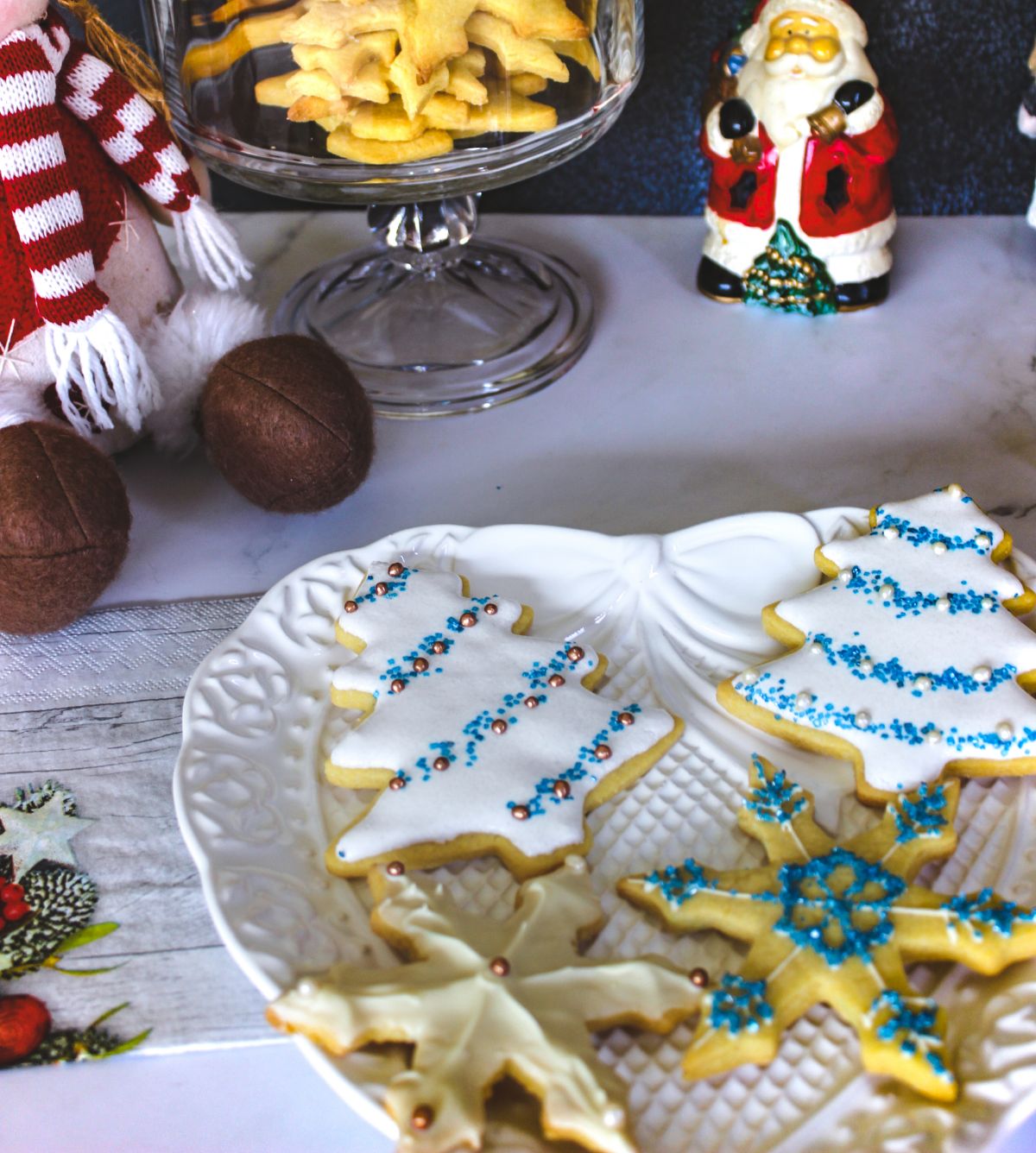 Christmas Sugar Cookies on a white serving plate on the bottom part of the image and the other bare cookies on the upper part of the image alongside with a Santa Claus miniature and stuffed toy.