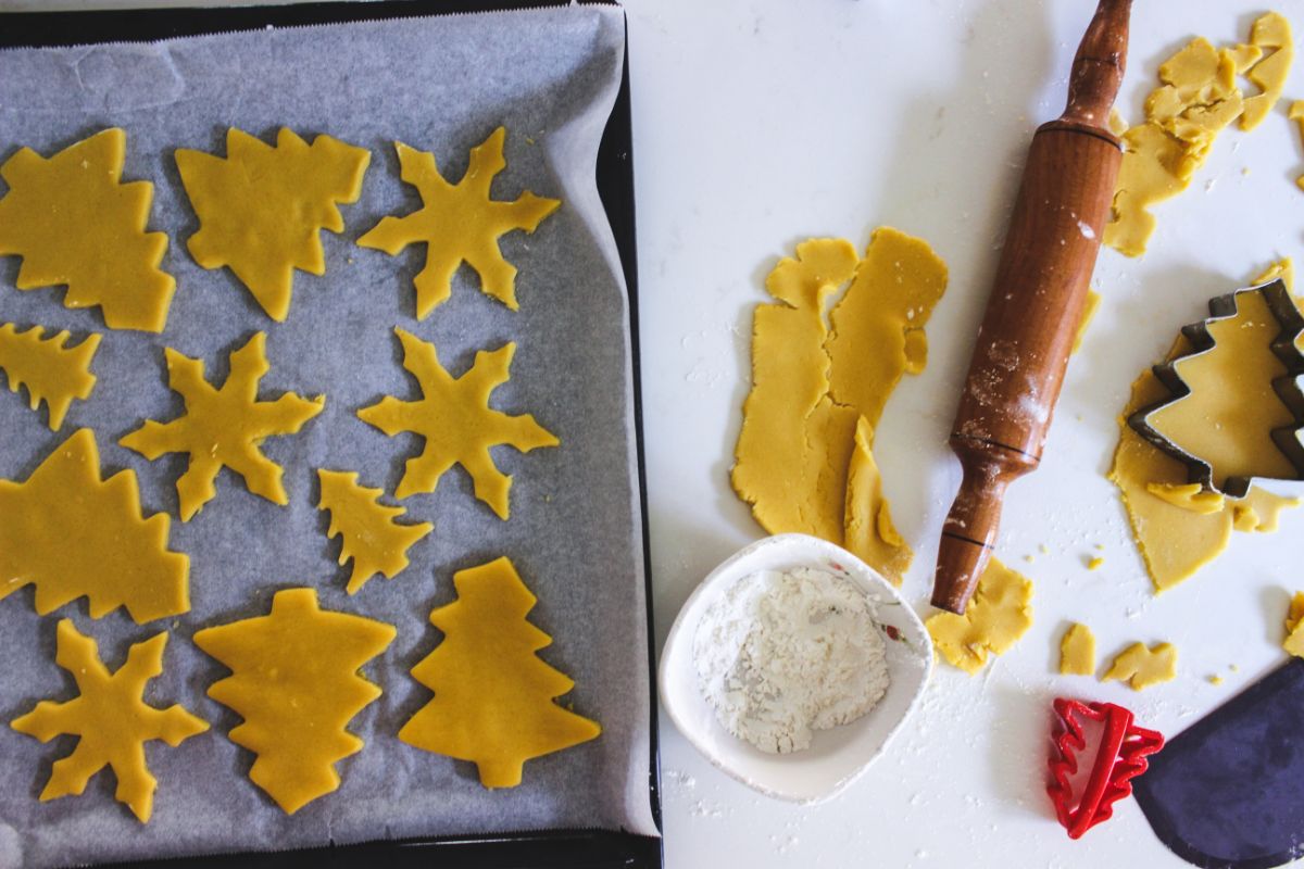 Cut outs of cookies on a parchment paper on the left part of the image and the remaining cookie dough being rolled out on the right side.