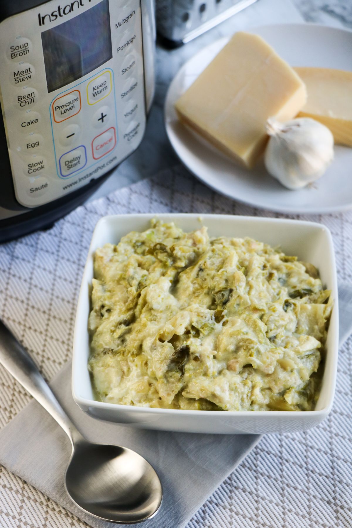 A vertical image of Instant Pot Creamed Brussel Sprouts in a white serving bowl with blocks of parmesan, garlic, and the Instant Pot in the background.